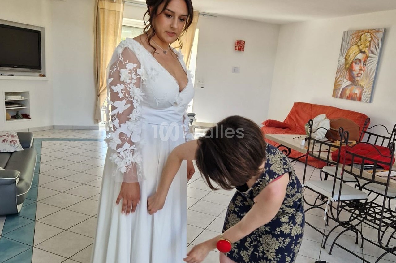 Une femme ajuste la robe blanche d'une mariée dans un salon lumineux avec des meubles modernes.