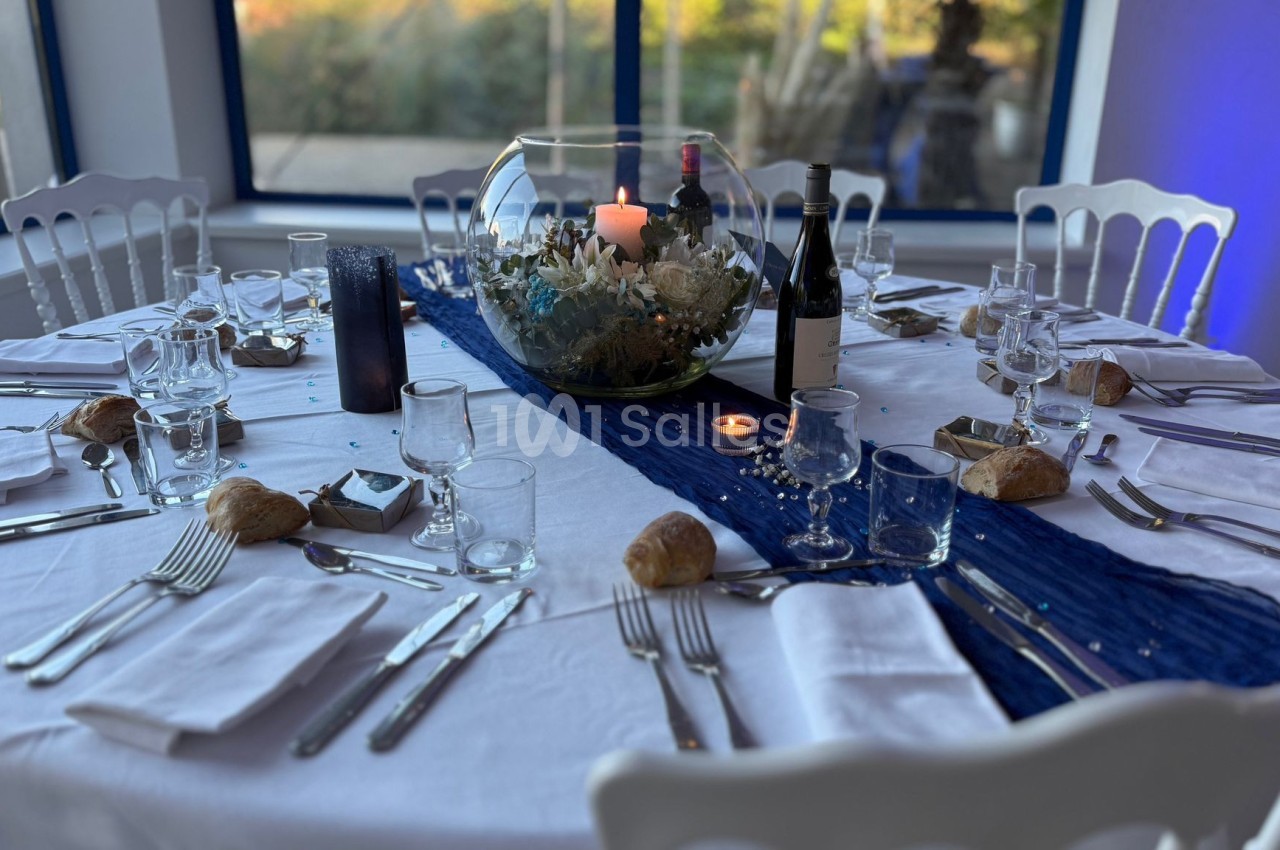 Table ronde dressée avec nappes blanches, décoration marine, verres, couverts, bougies et bouteilles de vin.