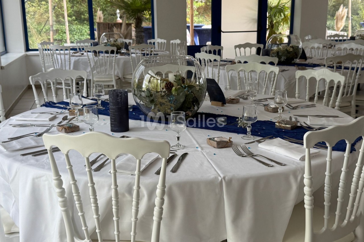 Salle de réception avec tables rondes dressées, nappes blanches, chaises blanches et centres de table décoratifs.