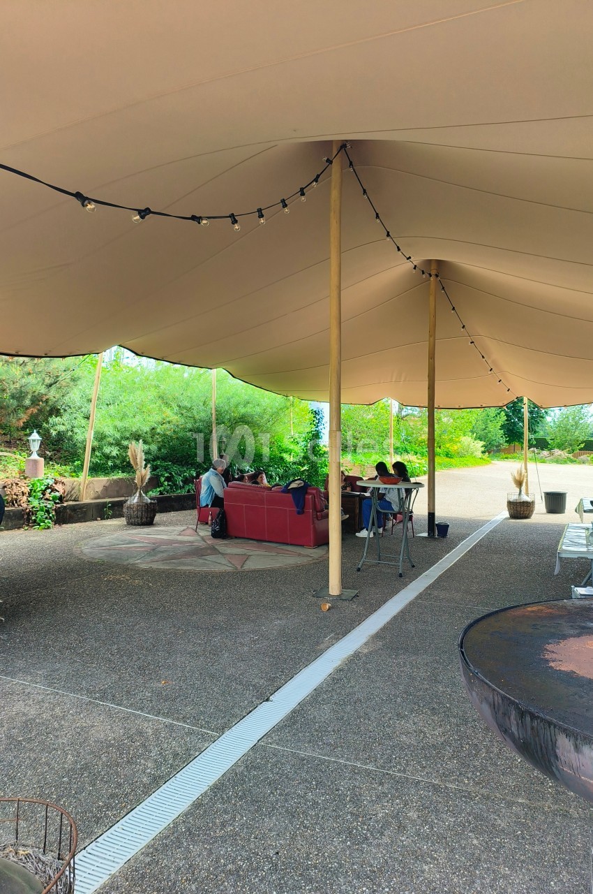Espace extérieur couvert par une tente beige, avec des tables et des personnes assises dans un cadre verdoyant.