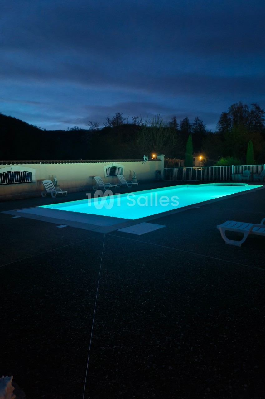 Piscine extérieure éclairée de nuit, entourée de chaises longues et d'arbres en arrière-plan.