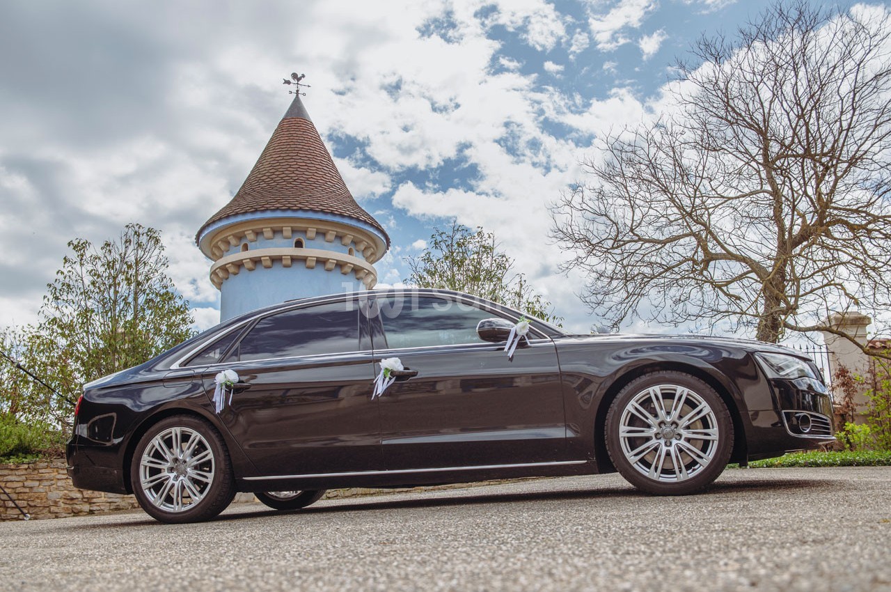 Voiture noire de luxe stationnée devant une tour en pierre avec un toit conique, sous un ciel partiellement nuageux.