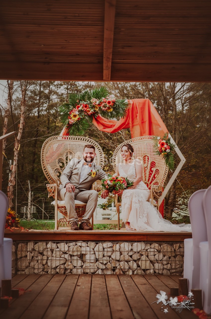 Un couple assis sur des fauteuils en osier décorés de fleurs, sous une pergola ornée de tissus et compositions florales.