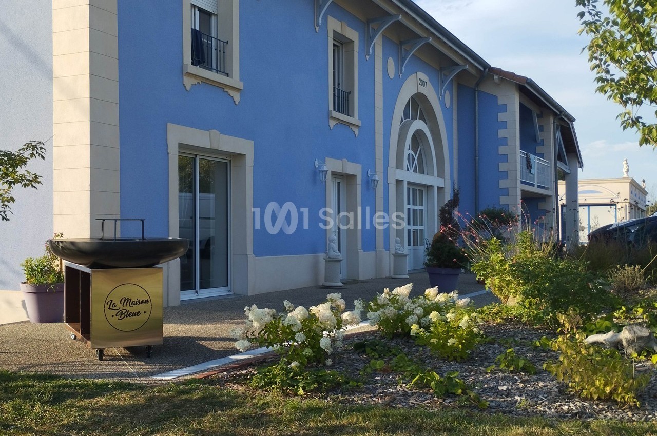 Façade d'un bâtiment bleu avec des fenêtres, un parterre fleuri et un panneau indiquant ’La Maison Rose’.