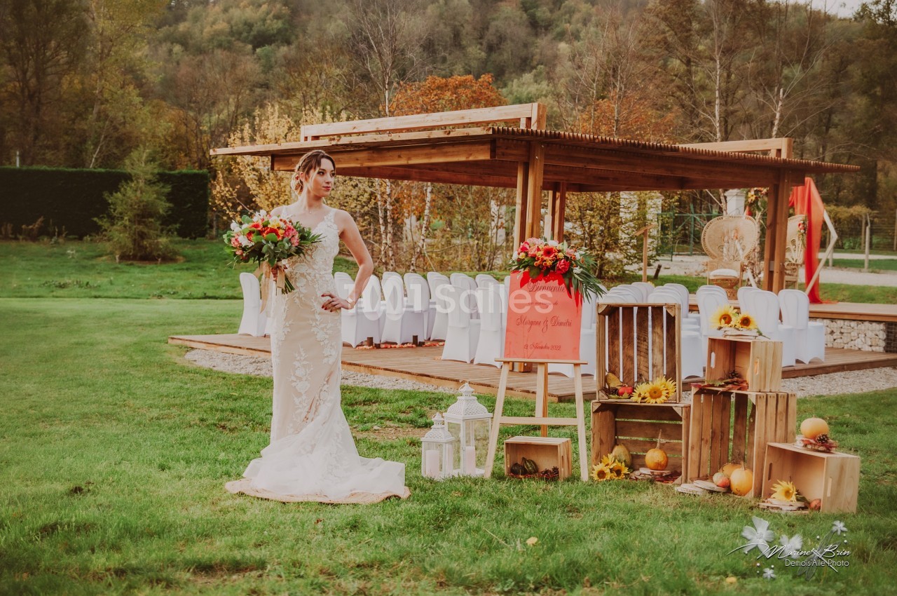 Une mariée en robe blanche tient un bouquet devant une cérémonie en extérieur, décorée de fleurs et de citrouilles.