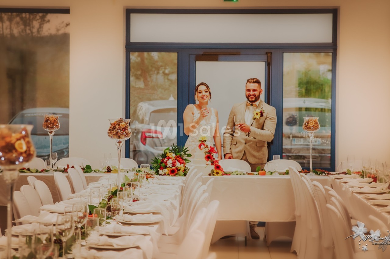 Un couple en tenue de mariage sourit debout derrière une table décorée dans une salle de réception.