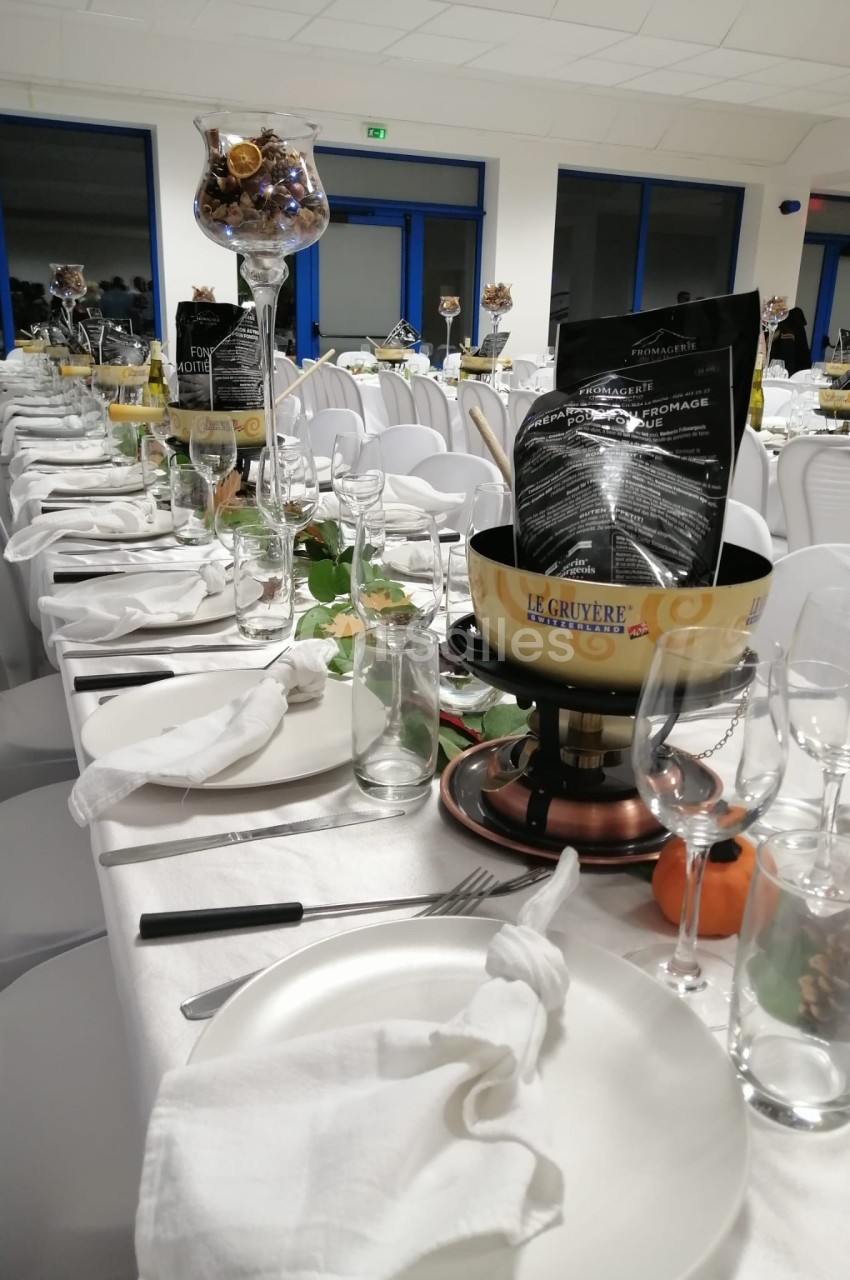Tables dressées pour un repas, avec nappes blanches, assiettes, verres, décorations et un caquelon à fondue au premier plan.