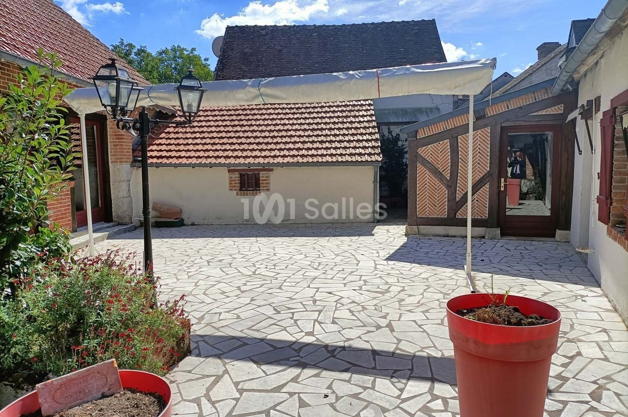 Cour pavée avec des bâtiments aux toits en tuiles, pots de fleurs rouges au premier plan et ciel dégagé en arrière-plan.