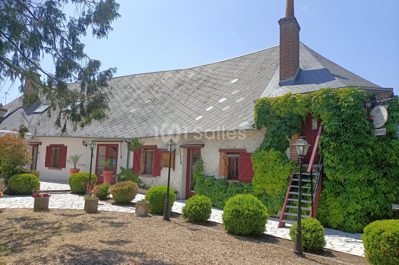 Maison traditionnelle avec façade blanche, volets rouges, toit en ardoise et vigne grimpante, entourée de végétation.