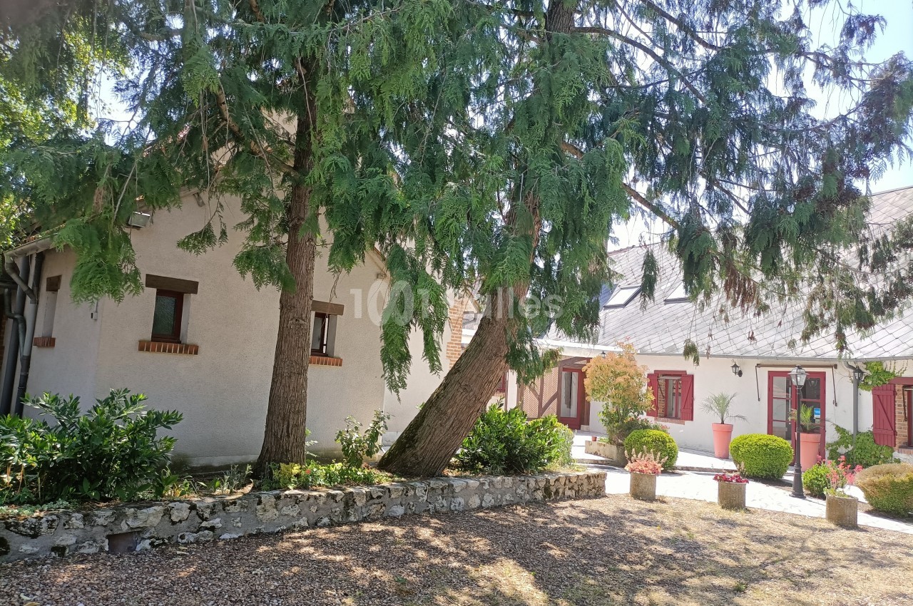 Cour d'une maison avec façade blanche, volets rouges, arbres imposants et jardinières fleuries sous un ciel dégagé.