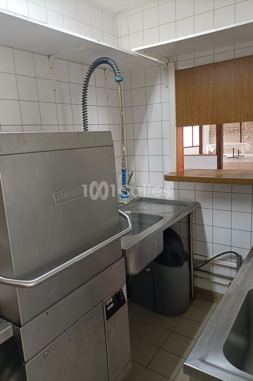 Lave-vaisselle industriel en inox dans une cuisine carrelée blanche, avec un évier et un robinet flexible à proximité.