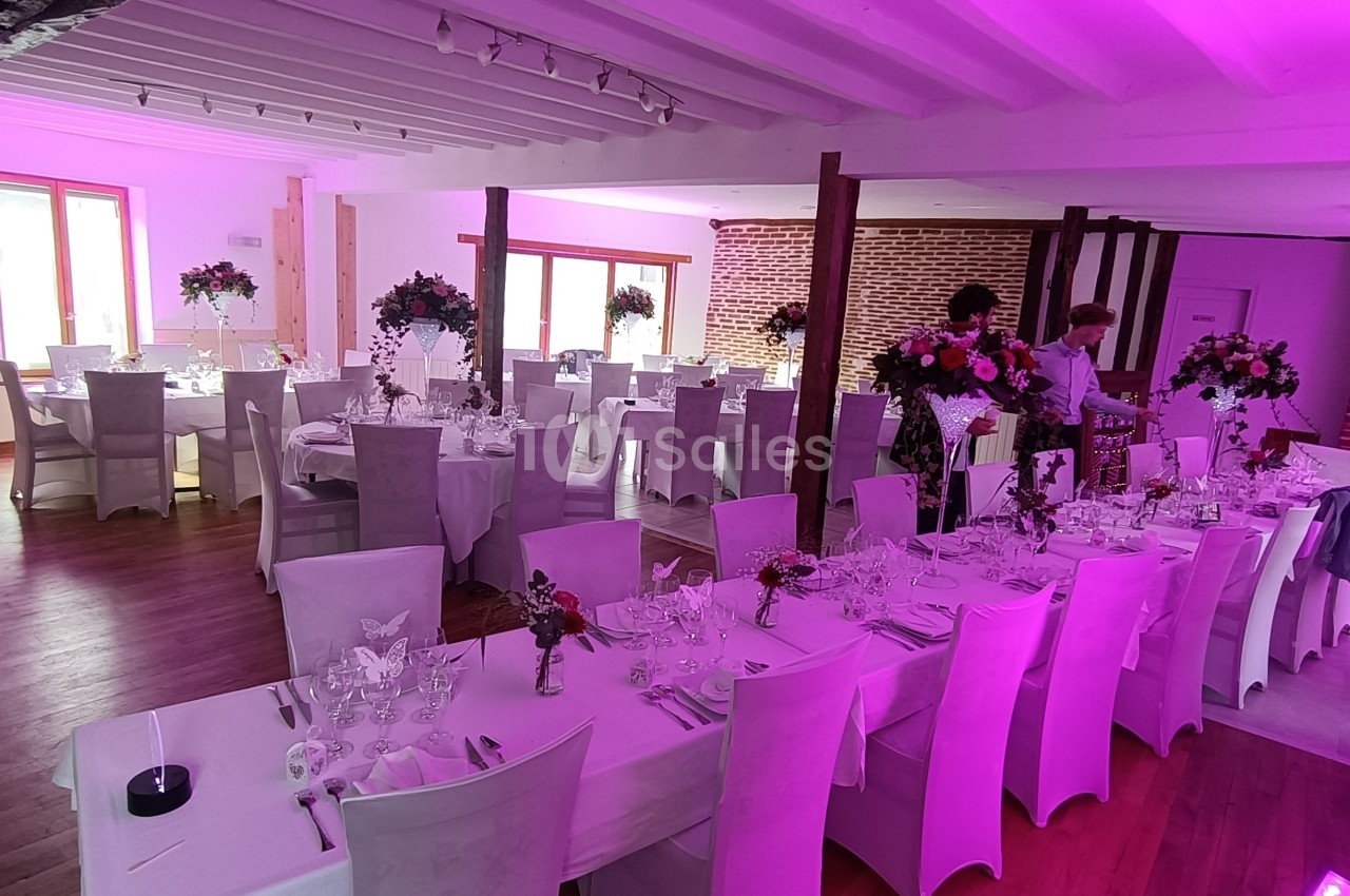 Salle de réception décorée avec des tables dressées, nappes blanches, chaises assorties et éclairage rose.