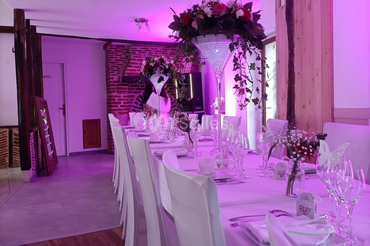 Salle de réception décorée avec une longue table dressée, nappes blanches, fleurs et éclairage rose tamisé.