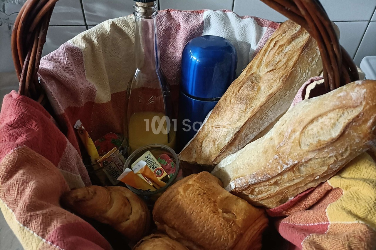 Panier en osier contenant des baguettes, viennoiseries, jus de fruit, thermos et confitures, posé sur une table.