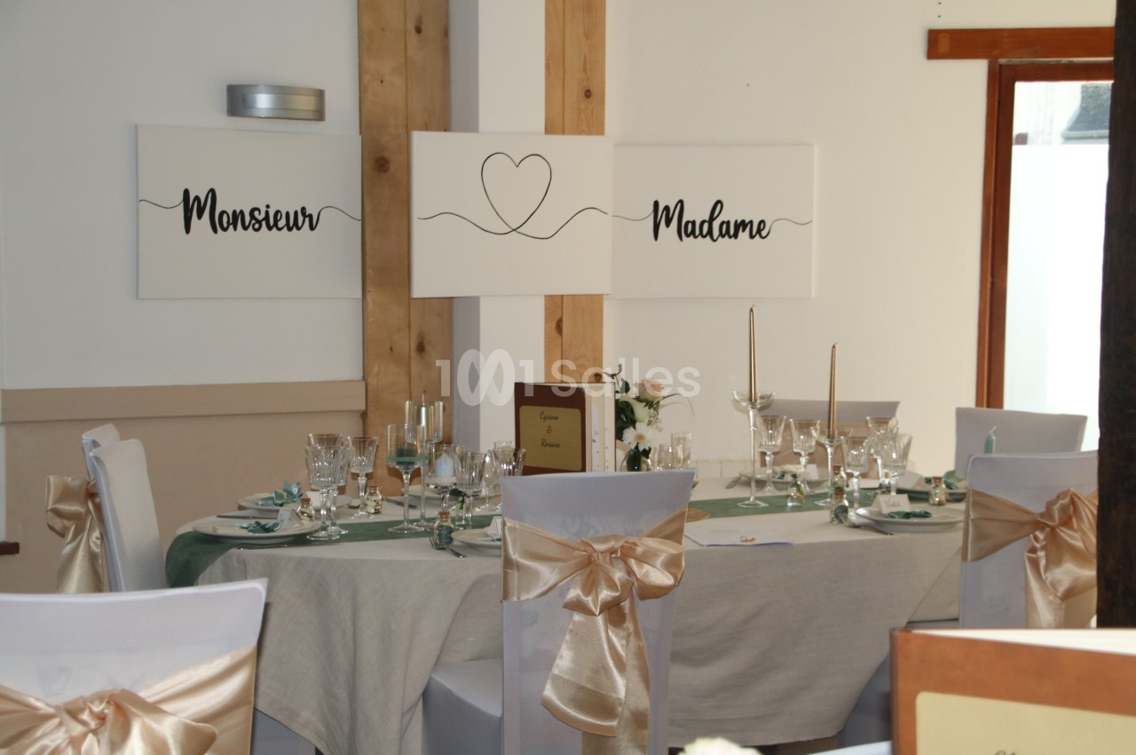 Table de réception décorée avec nappes claires, chaises ornées de nœuds dorés et panneaux ’Monsieur’ et ’Madame’ au mur.