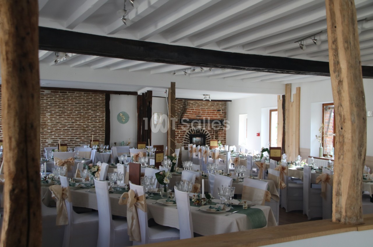 Salle de réception décorée avec des tables dressées, des chaises blanches et des éléments rustiques en bois et briques.