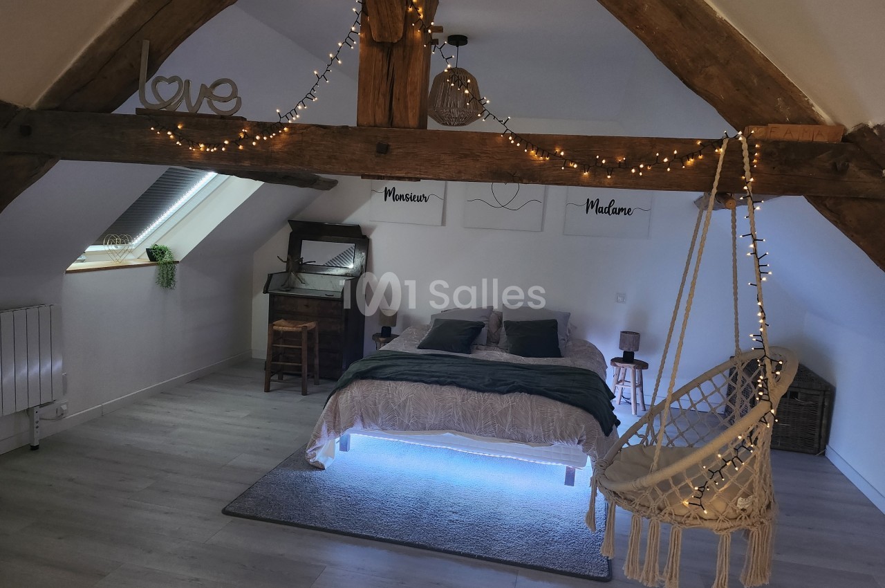 Chambre mansardée avec poutres apparentes, lit double, fauteuil suspendu et guirlandes lumineuses décoratives.