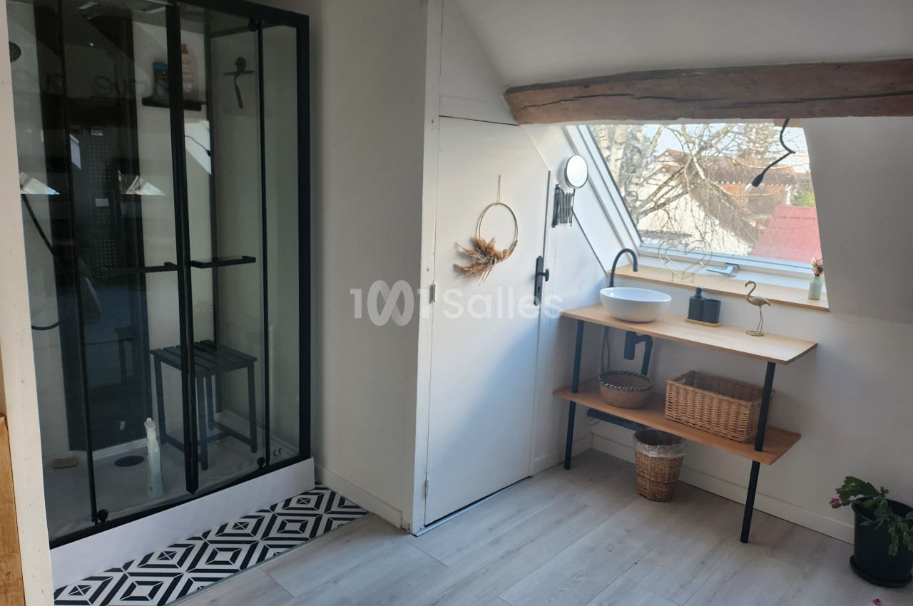Salle de bain lumineuse sous combles avec douche vitrée, lavabo sur meuble en bois et fenêtre donnant sur l'extérieur.
