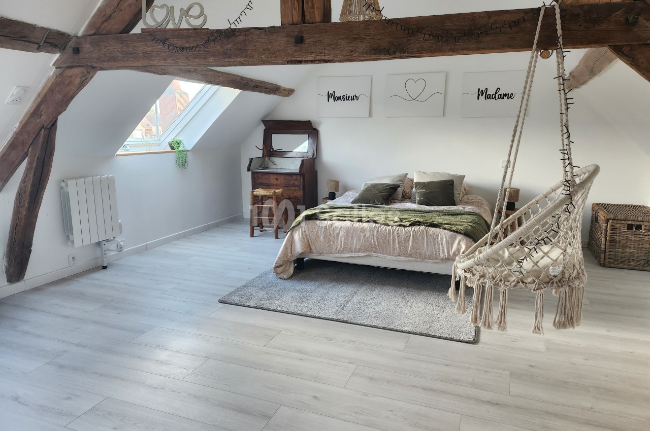 Chambre lumineuse sous combles avec poutres apparentes, lit double, fauteuil suspendu et bureau en bois.