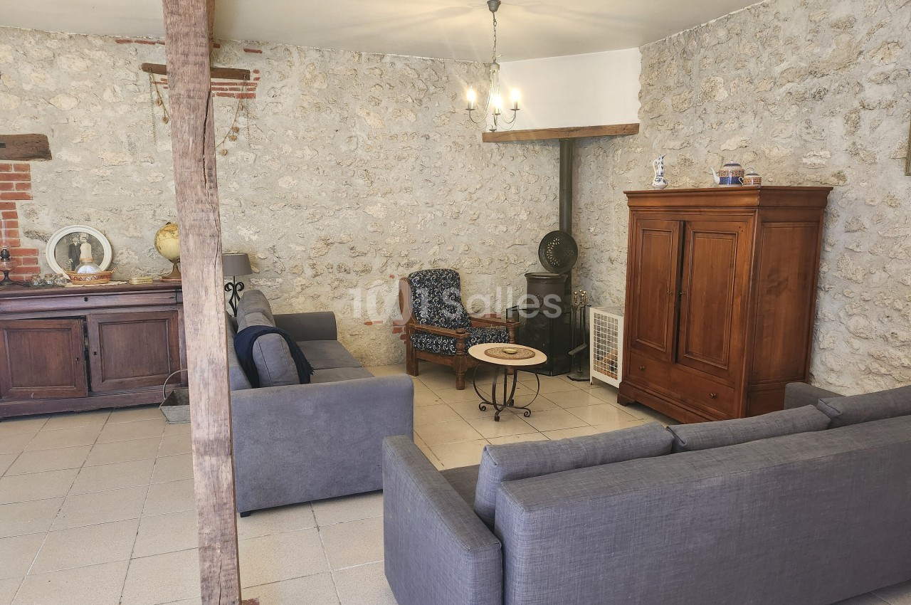 Salon avec murs en pierre, canapés gris, fauteuil, armoire en bois, table basse et éclairage suspendu.
