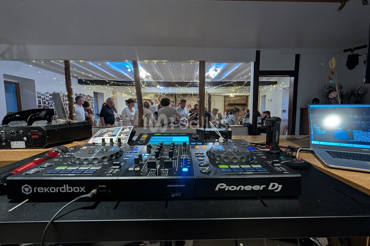 Vue d'une table de mixage DJ avec des invités en arrière-plan dans une salle décorée de guirlandes lumineuses.
