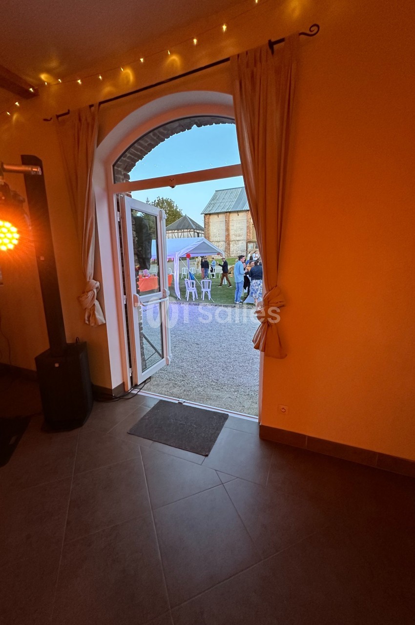 Vue depuis une salle éclairée vers une porte ouverte donnant sur un jardin avec des personnes et des tables en extérieur.