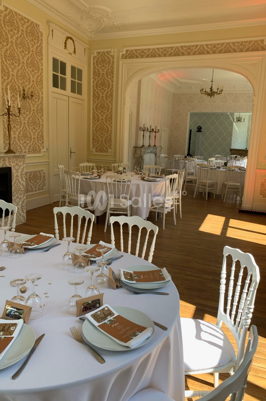 Salle élégante avec tables rondes dressées pour un événement, chaises blanches et lumière naturelle traversant les fenêtres.