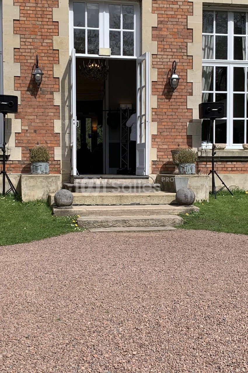 Entrée d'un bâtiment en briques avec porte ouverte, escaliers, pots de fleurs et haut-parleurs disposés de chaque côté.