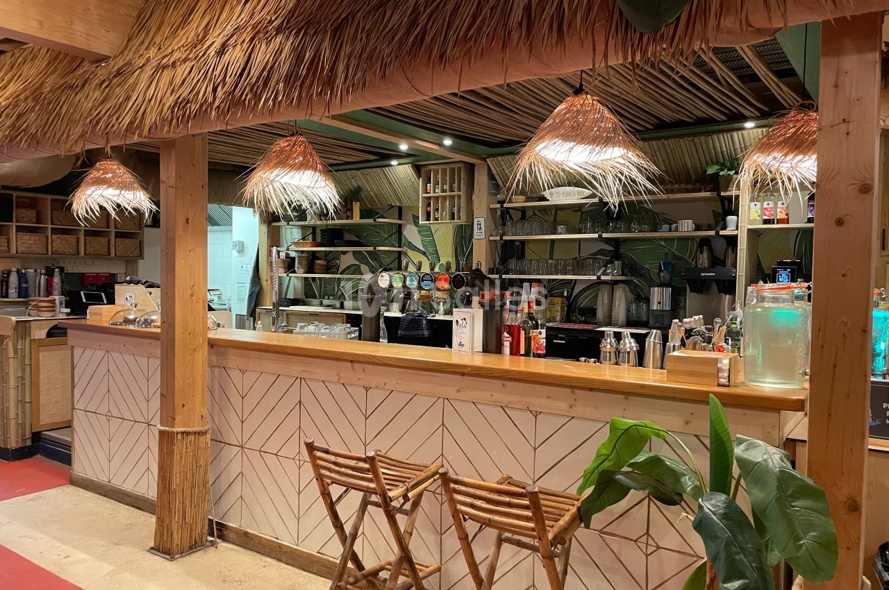 Comptoir en bois d'un bar à thème tropical avec tabourets pliants, éclairage en paille et décoration végétale.