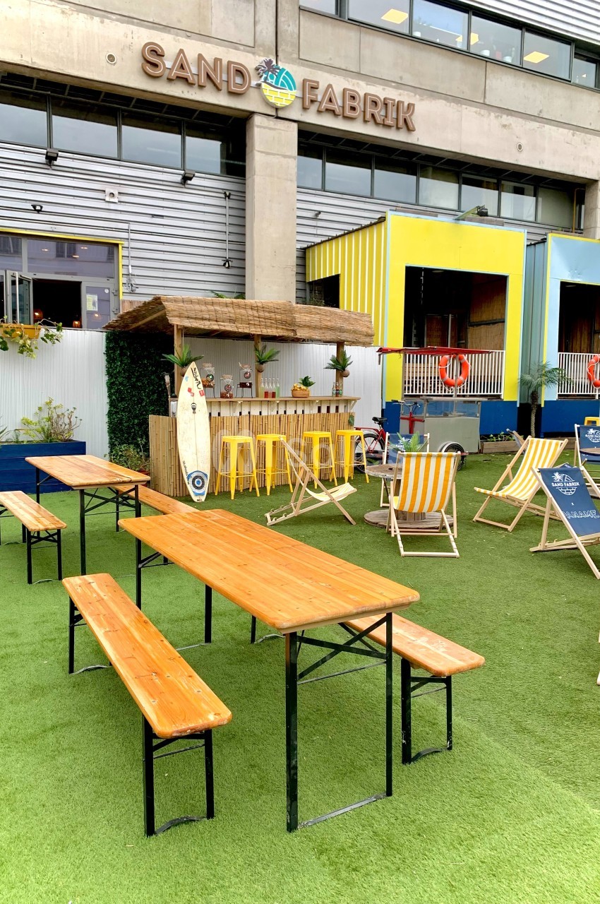 Tables en bois, chaises longues et bar en extérieur sur une terrasse avec gazon artificiel devant un bâtiment.