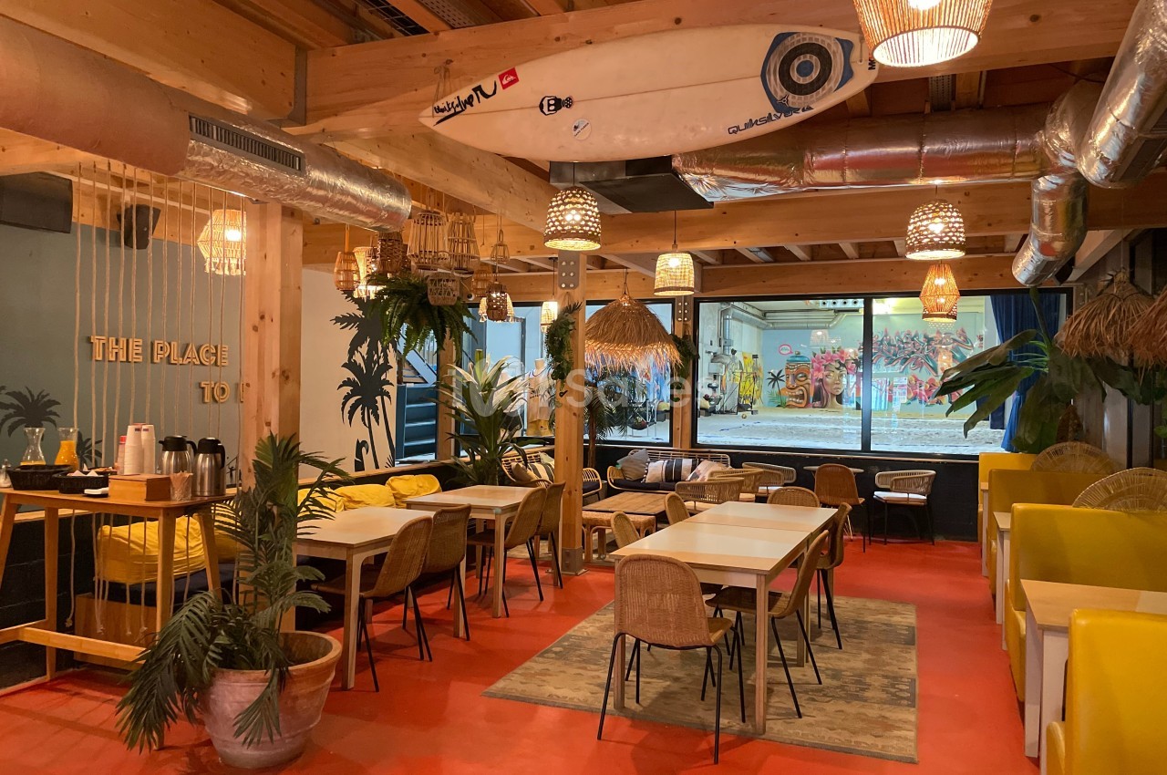Salle lumineuse avec tables en bois, chaises en rotin, décor tropical et planche de surf suspendue au plafond.