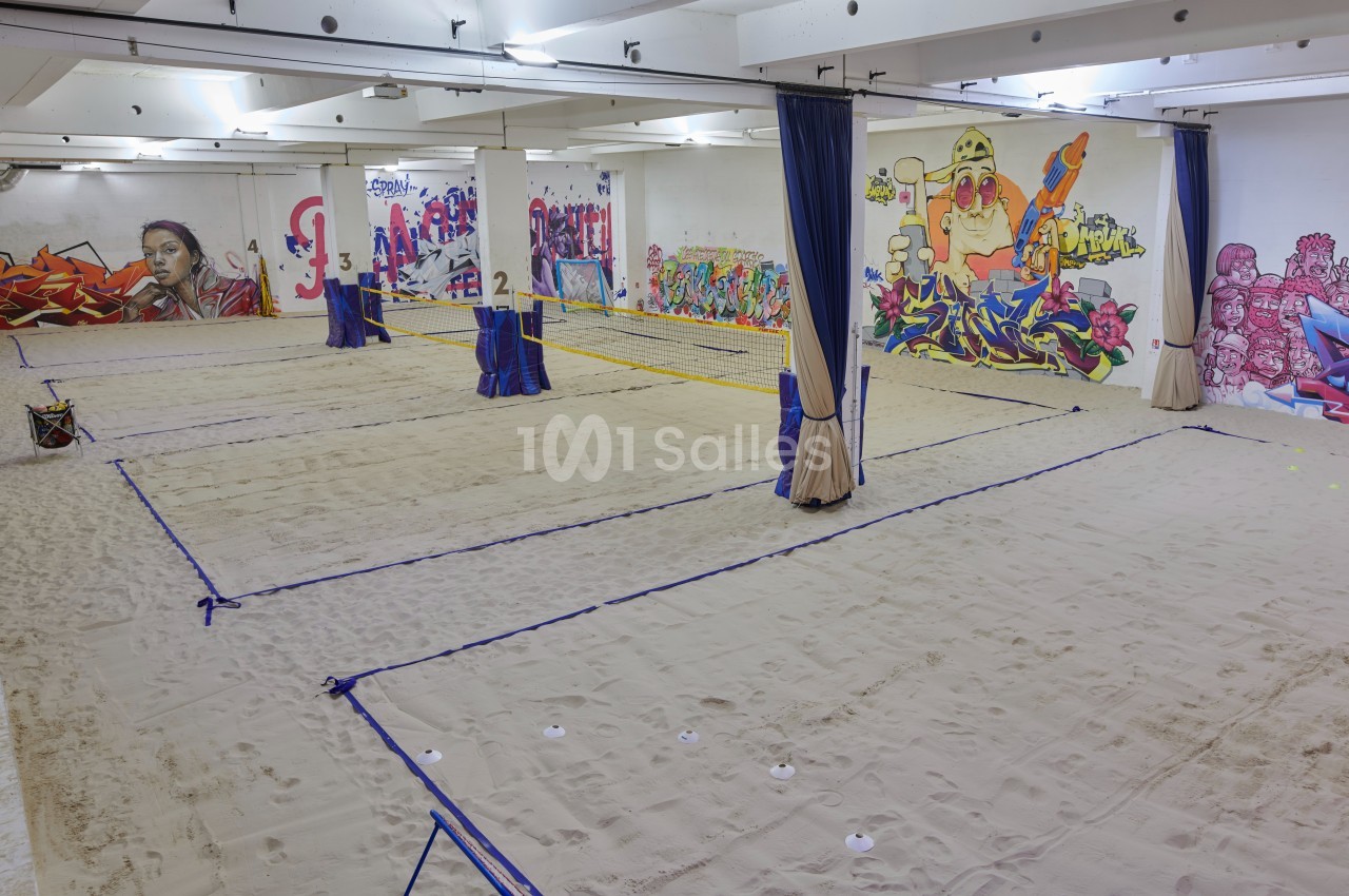 Terrain de beach-volley intérieur avec sol en sable, filets tendus et murs décorés de graffitis colorés.