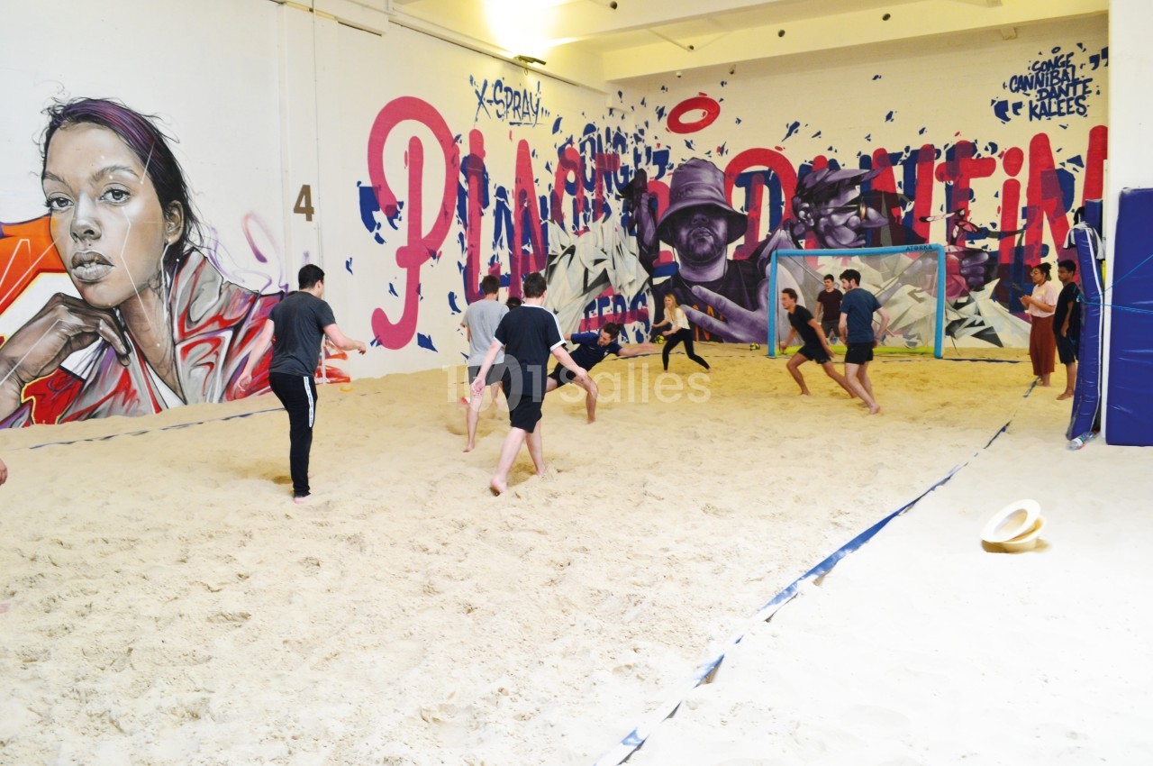 Des personnes jouent au football sur un terrain de sable intérieur, entouré de fresques murales colorées.