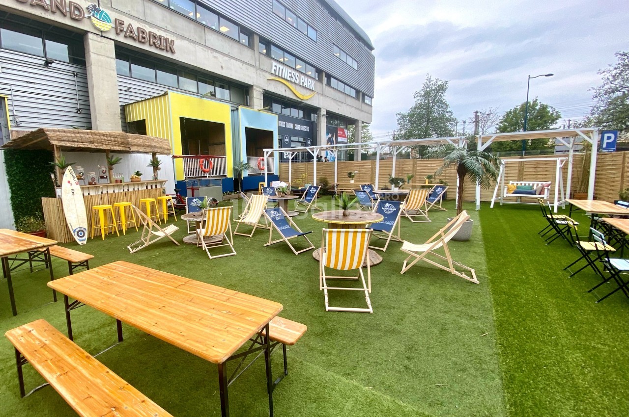 Espace extérieur avec chaises longues, tables en bois et gazon artificiel, entouré de bâtiments modernes.