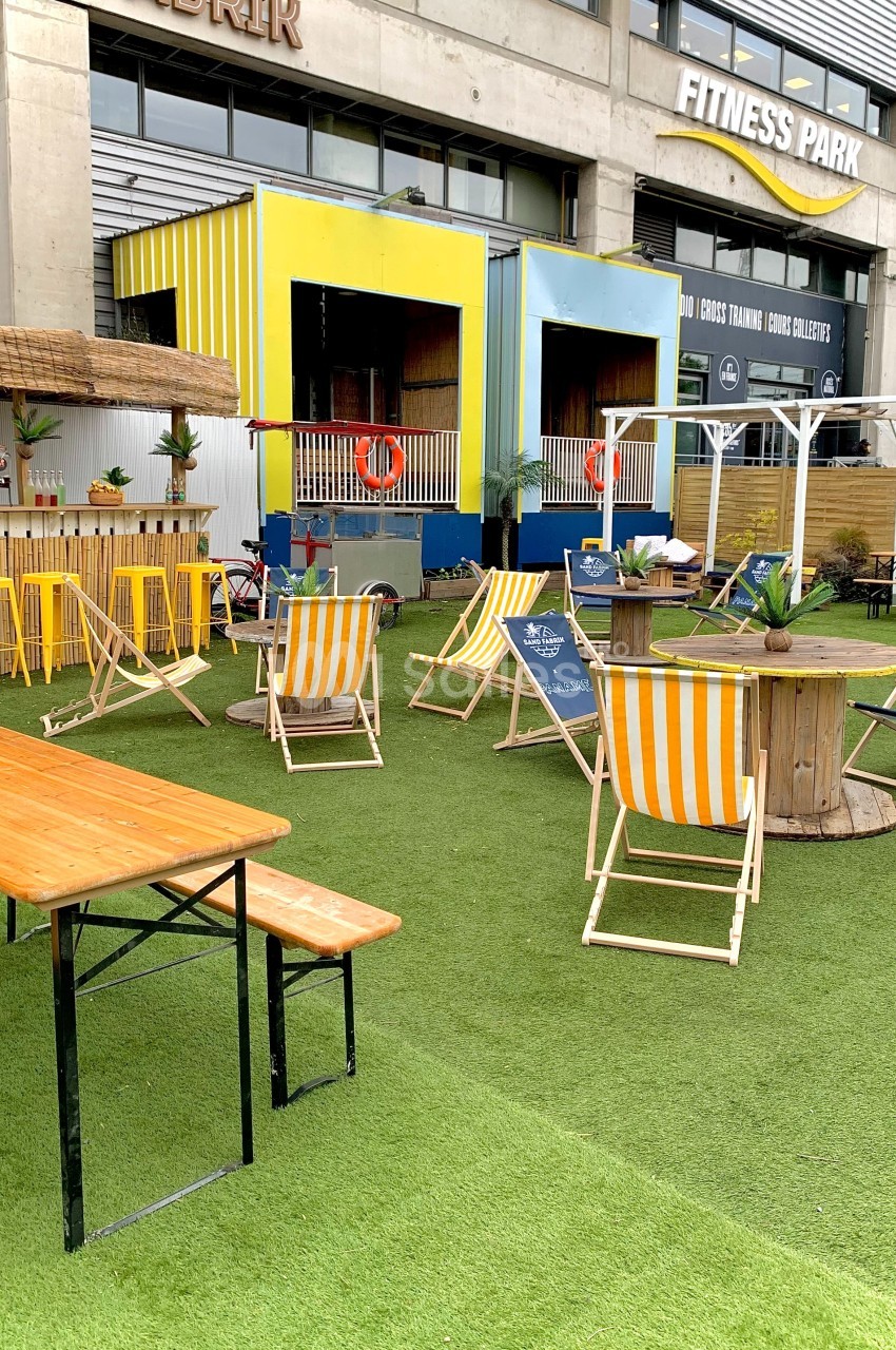 Terrasse extérieure avec chaises longues, tables en bois et décor tropical sur un sol en gazon synthétique.