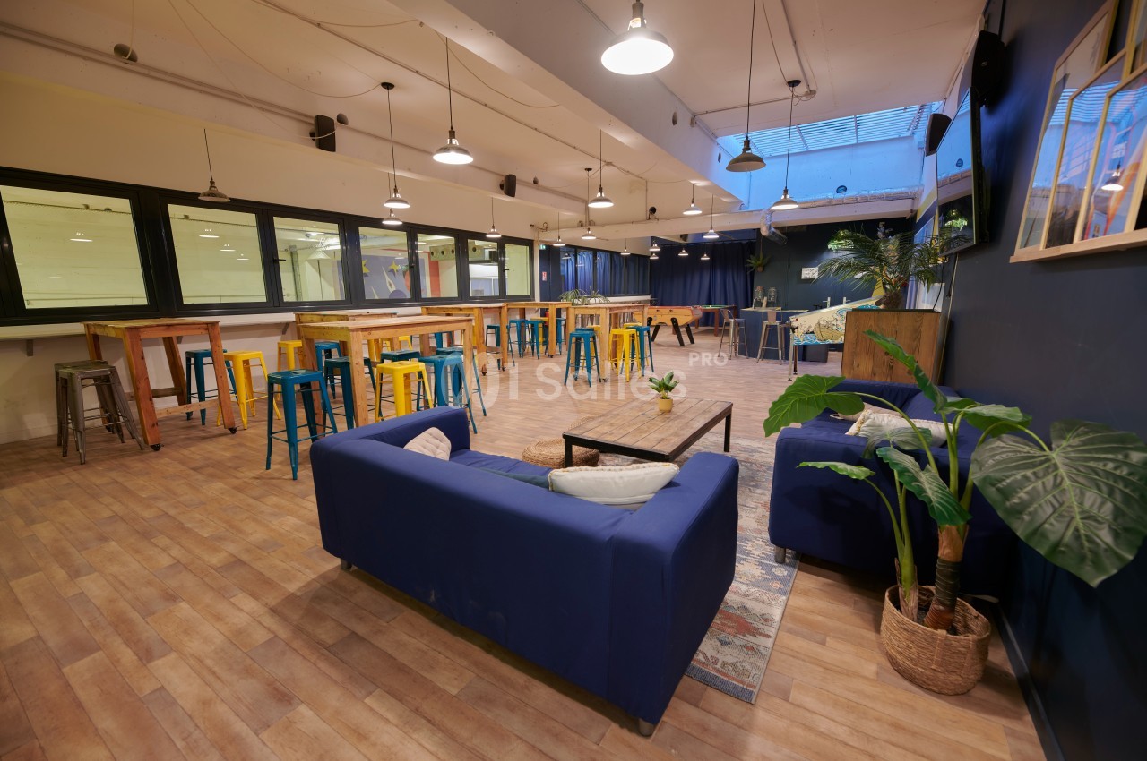 Salle spacieuse avec canapés bleus, tables hautes en bois, tabourets colorés et grandes fenêtres vitrées.