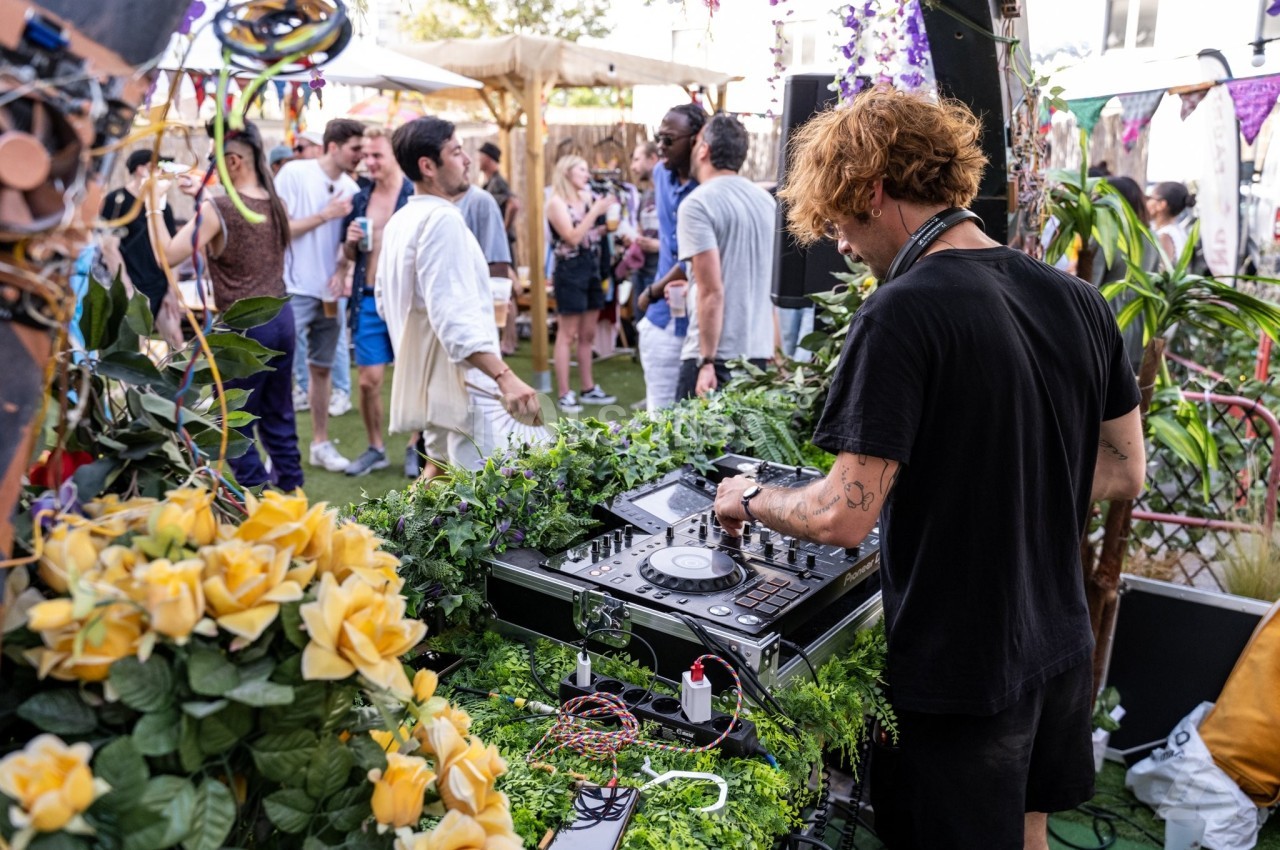Un DJ mixe en plein air lors d'un événement festif, entouré de plantes et de participants discutant en arrière-plan.