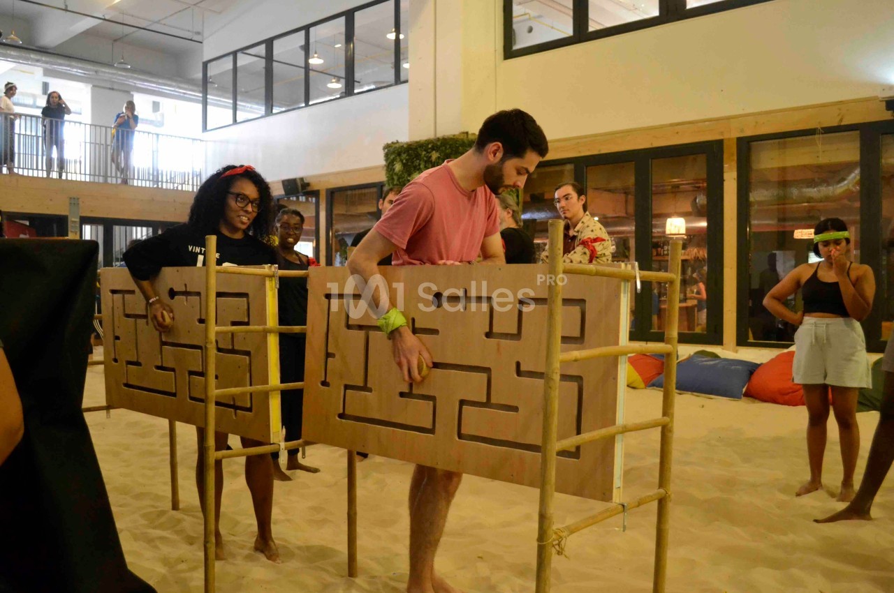 Des personnes participent à un jeu d'adresse avec des labyrinthes en bois dans un espace intérieur sur sol sablé.