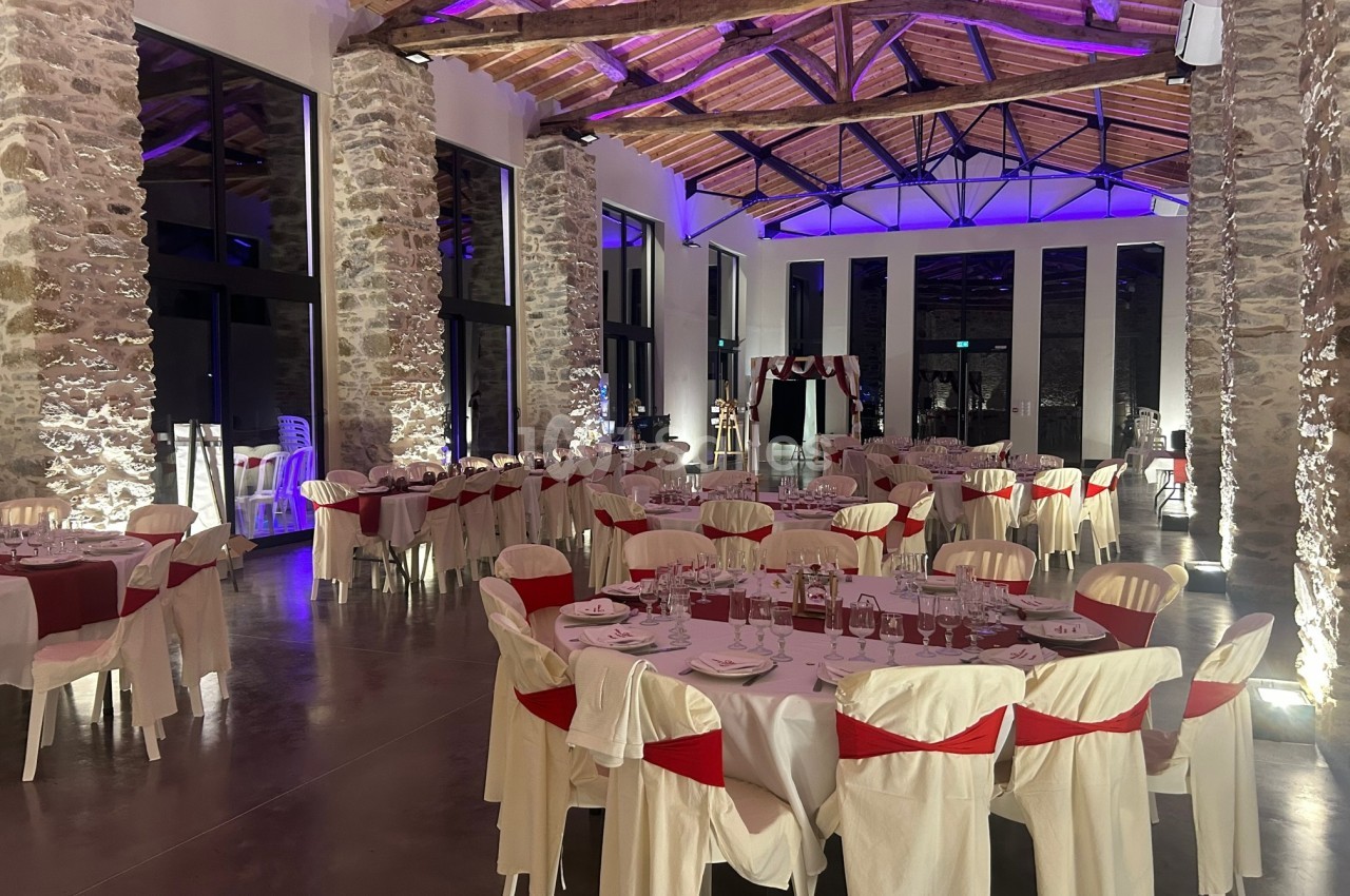 Salle de réception décorée avec des tables rondes dressées, nappes blanches et chaises ornées de rubans rouges.
