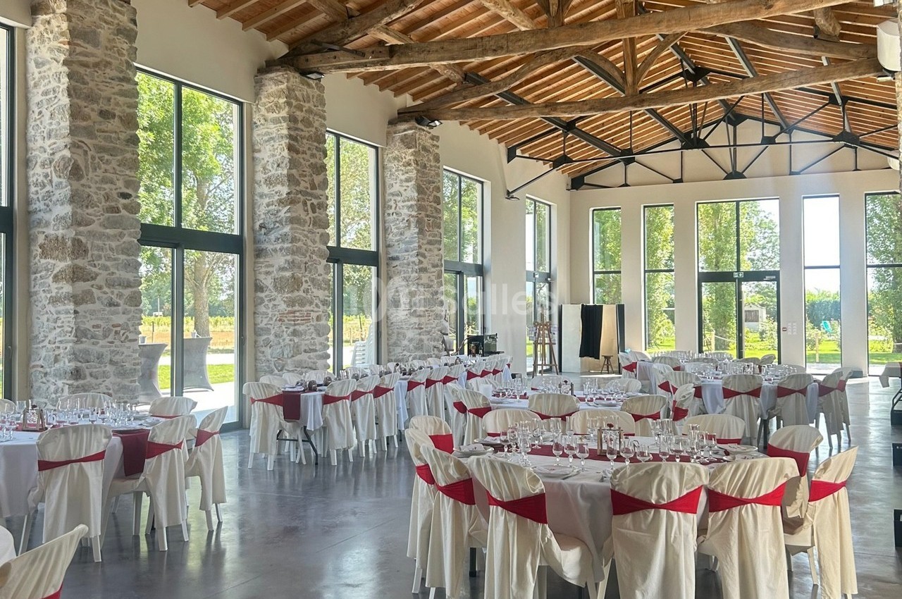 Salle de réception lumineuse avec de grandes baies vitrées, des murs en pierre et des tables dressées avec nappes blanches.