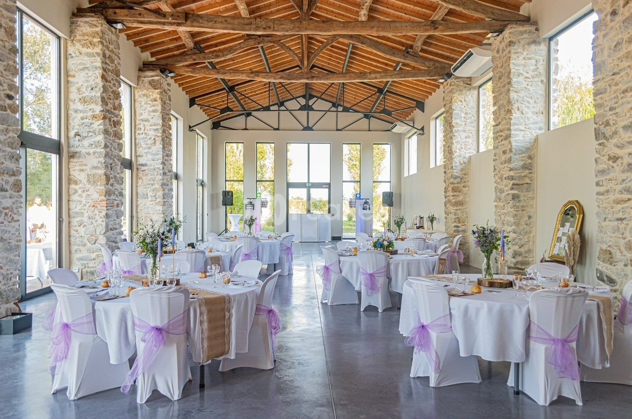 Salle de réception lumineuse avec des tables décorées de nappes blanches et rubans mauves, entourée de murs en pierre.