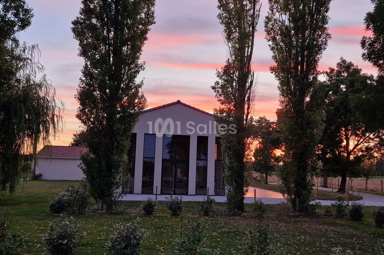Façade d'une maison moderne entourée d'arbres, au coucher du soleil avec un ciel aux teintes roses et orangées.