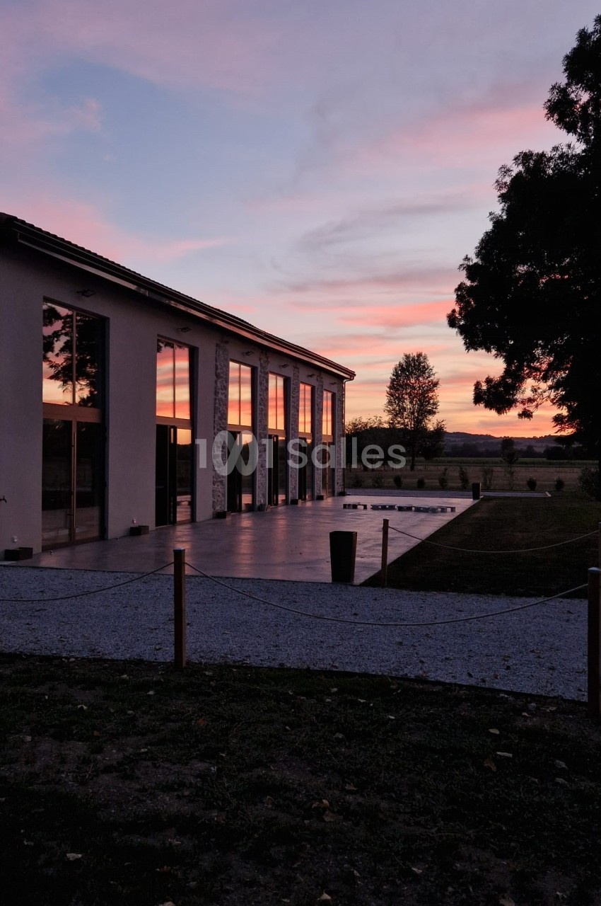 Bâtiment moderne avec de grandes baies vitrées reflétant un ciel rose au coucher du soleil, entouré d'arbres et d'un jardin.