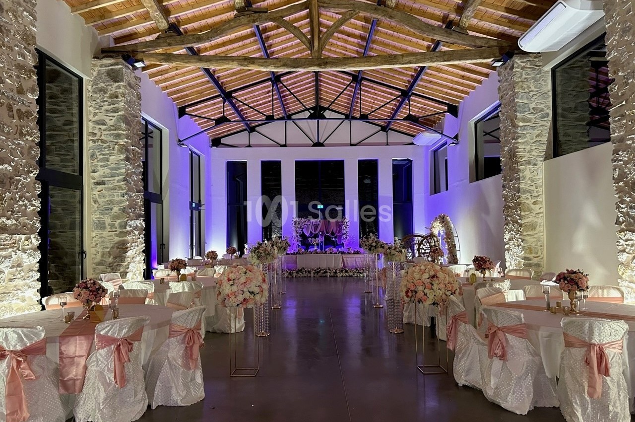 Salle de réception décorée avec des tables rondes ornées de nappes blanches et roses, éclairage chaleureux et arc floral.