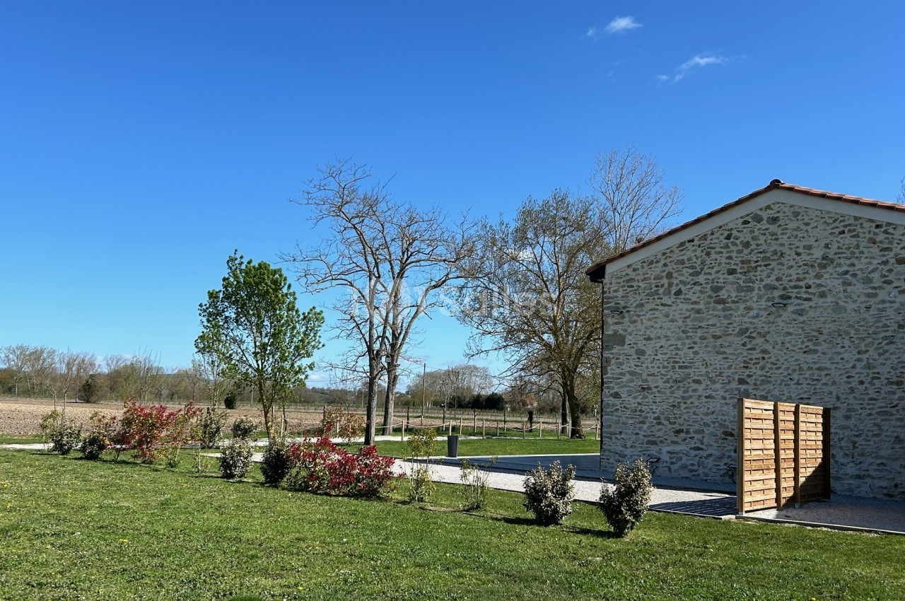 Jardin avec pelouse, arbustes et arbres, bordé par un bâtiment en pierre sous un ciel bleu dégagé.