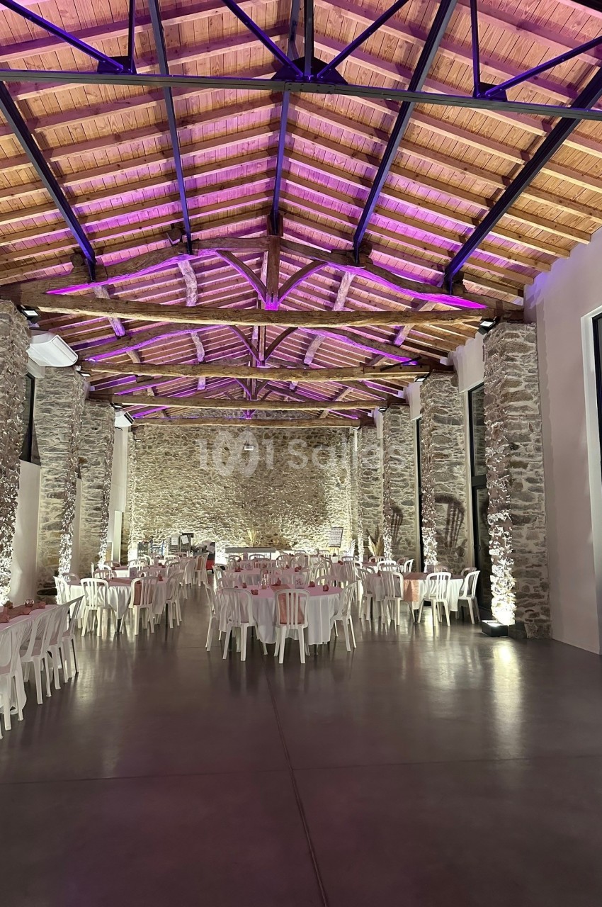 Grande salle en pierre avec plafond en bois, éclairage violet et tables blanches disposées pour un événement.