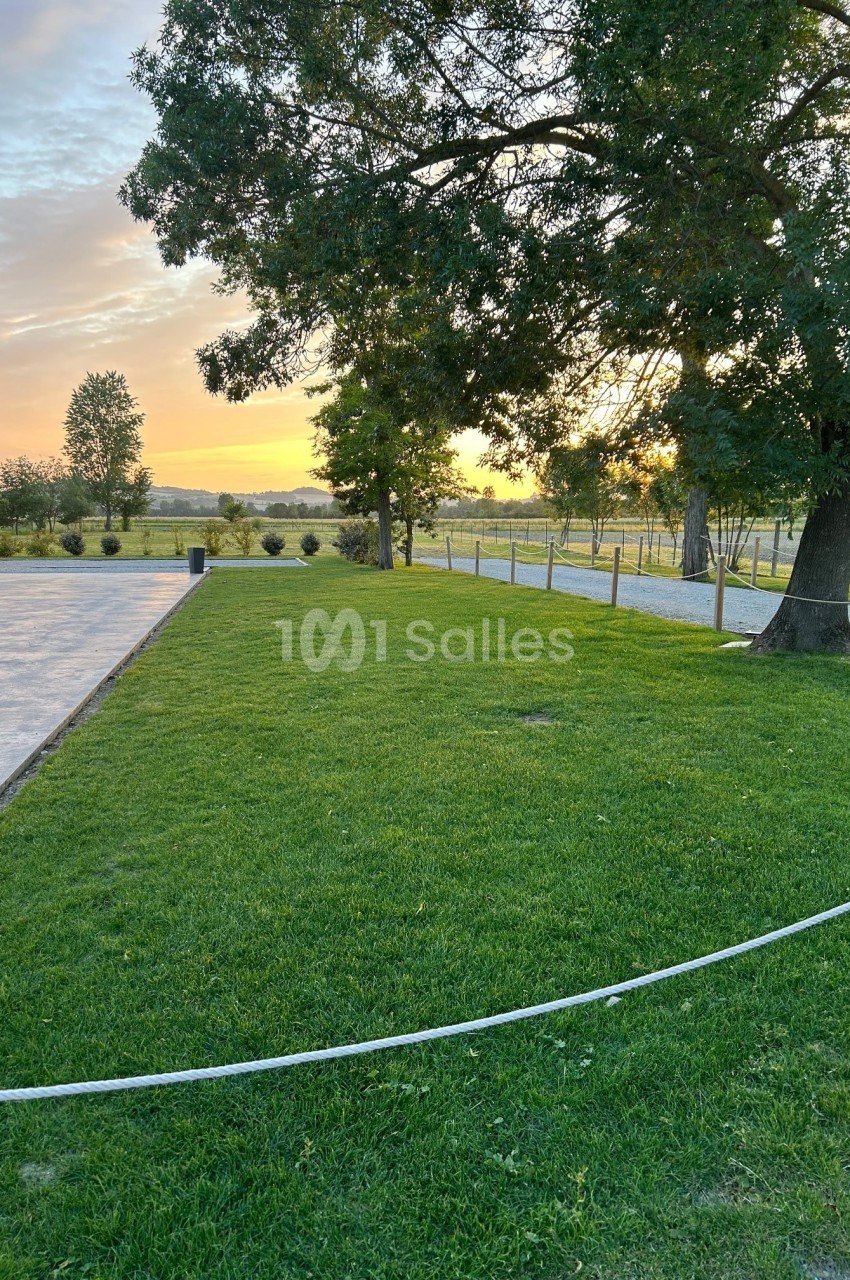 Pelouse verte bordée d'arbres au coucher du soleil, avec une terrasse à gauche et une clôture en arrière-plan.