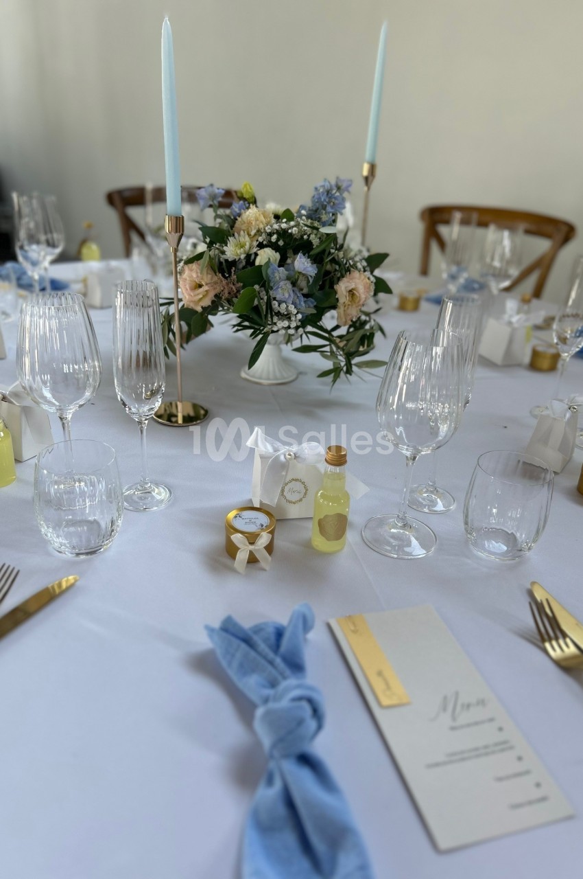 Table élégamment dressée avec des couverts dorés, serviettes bleues, bougies, fleurs et menus pour un repas formel.
