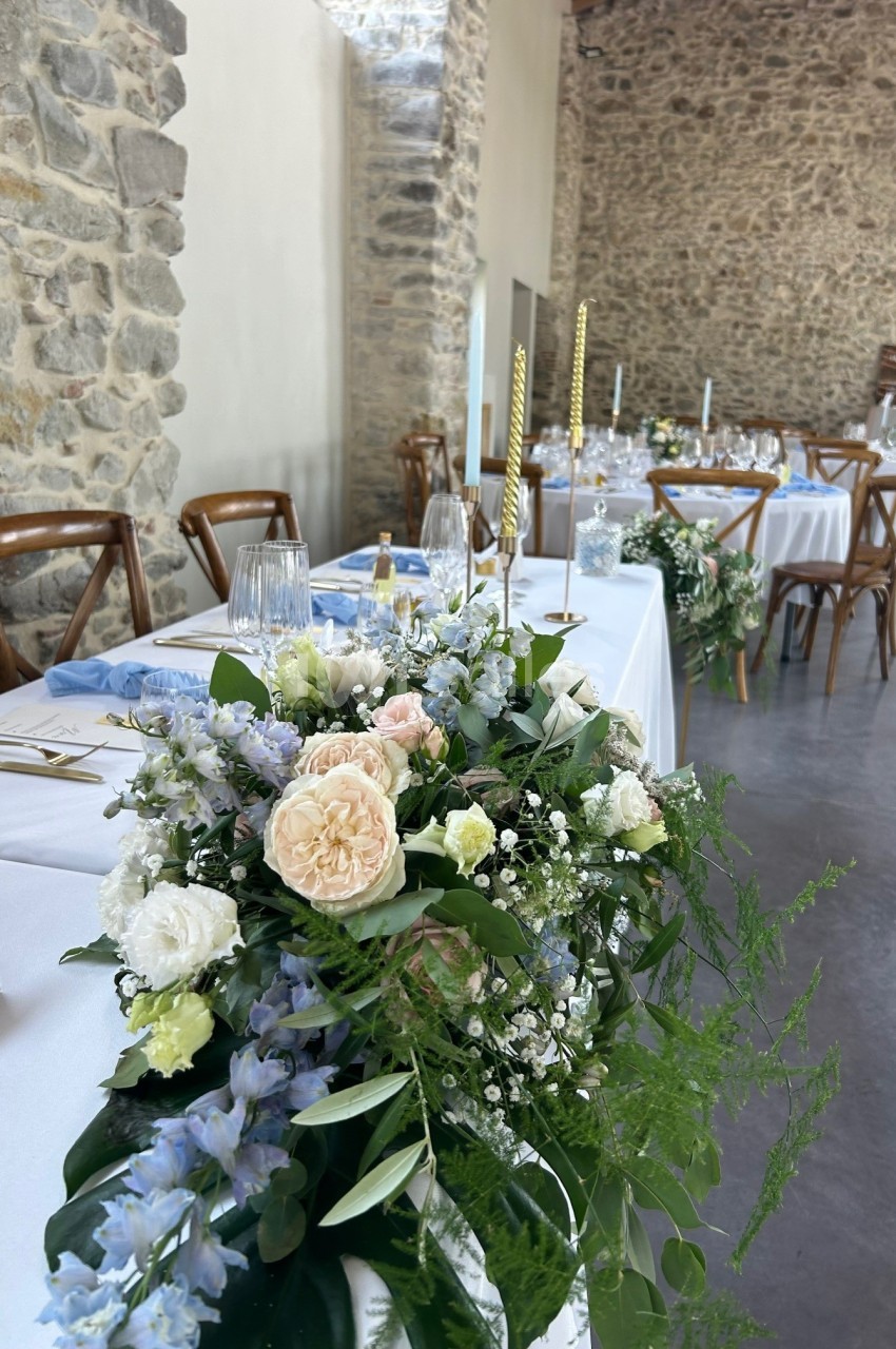 Décoration florale avec roses, gypsophiles et feuillage sur une table de réception dans une salle en pierre.