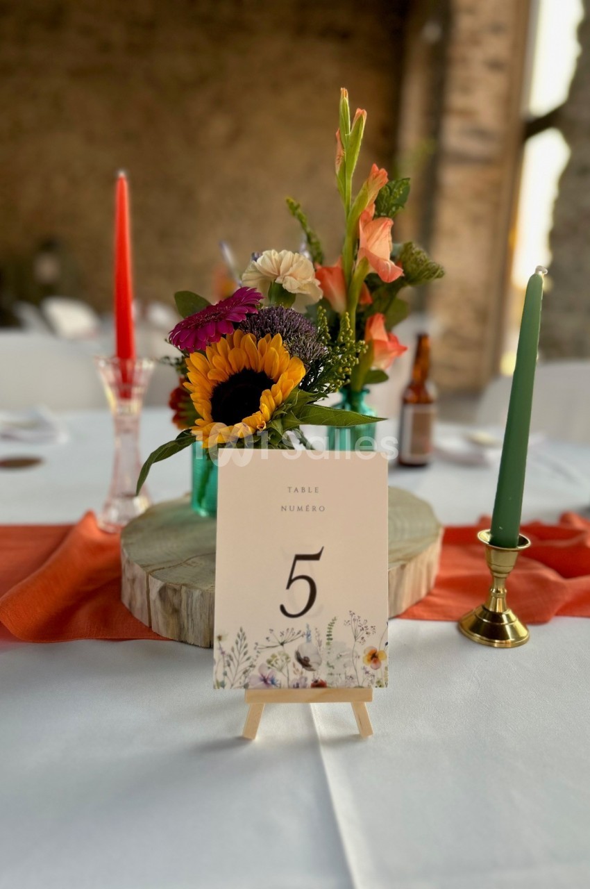 Centre de table avec fleurs colorées, bougies rouge et verte, et une carte indiquant ’Table numéro 5’.